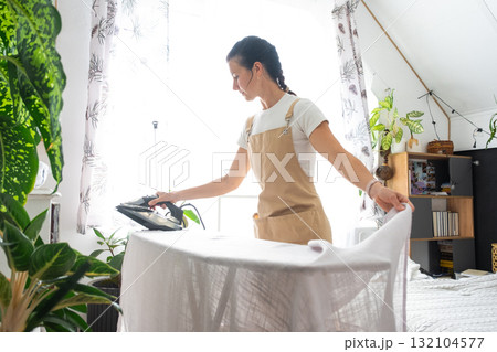 A housewife woman in an apron irons linen curtains in a room on an ironing board A housewife woman in an apron irons linen curtains in a room on an ironing board 132104577