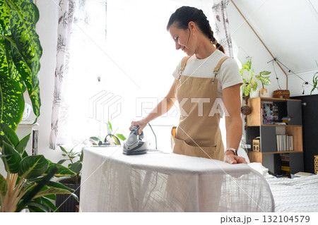 A housewife woman in an apron irons linen curtains in a room on an ironing board 132104579