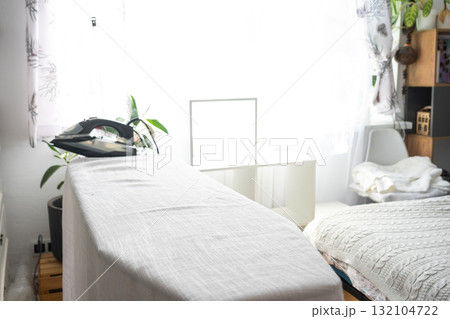 The iron is on the ironing board with a plain cloth in the room. The iron is on the ironing board with a plain cloth in the room. 132104722