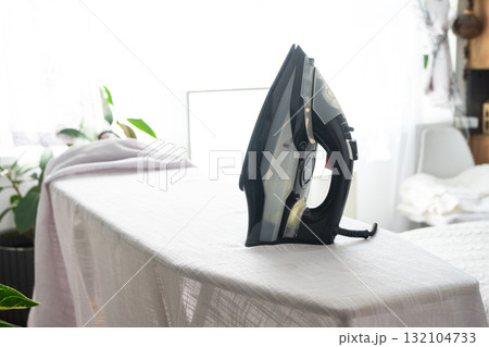 The iron is on the ironing board with a plain cloth in the room in close-up. The iron is on the ironing board with a plain cloth in the room in close-up. 132104733