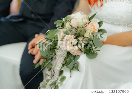 Close-up of a bride in white dress holding a romantic cascading bouquet with pastel roses, greenery, and lace ribbon 132107900