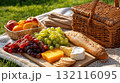 Picnic setup on grass with fruits, bread, cheese, and grapes beside a wicker basket 132116095