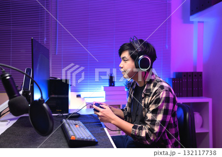 Young Asian gamer wearing headset playing video game at night with neon light, focused streaming setup and modern gaming lifestyle Young Asian gamer wearing headset playing video game at night with neon light, focused streaming setup and modern gaming lifestyle 132117738
