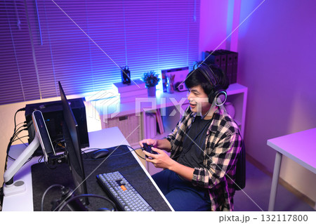 Young Asian gamer wearing headset playing video game at night with neon light, focused streaming setup and modern gaming lifestyle Young Asian gamer wearing headset playing video game at night with neon light, focused streaming setup and modern gaming lifestyle 132117800