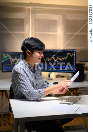 Young Asian trader analyzing financial charts on computer at night, focused on investment strategy 132117858