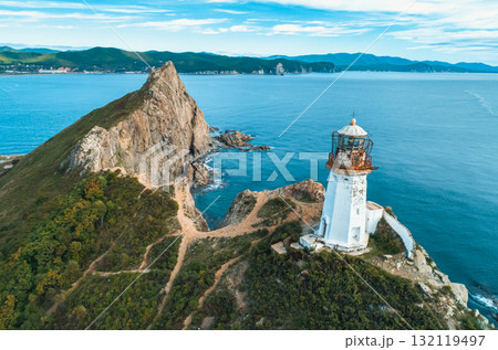 Lighthouse stands on rocky coastline, overlooking the ocean with mountains in the background during a clear day Lighthouse stands on rocky coastline, overlooking the ocean with mountains in the background during a clear day 132119497