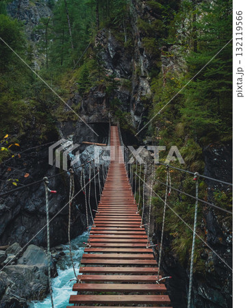 Wooden suspension bridge stretches over a rocky gorge surrounded by lush green trees in the mountains 132119566