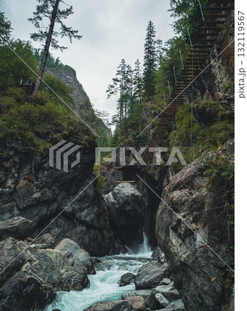 Water rushes over smooth stones at the base of rugged cliffs, while a wooden bridge stretches across the rocky landscape. 132119567