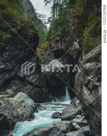 Water rushes over smooth stones at the base of rugged cliffs, while a wooden bridge stretches across the rocky landscape. 132119568