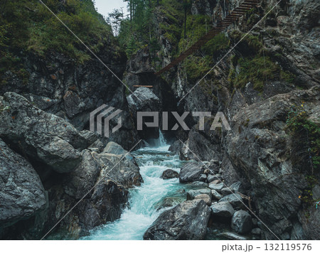 Water rushes over smooth stones at the base of rugged cliffs, while a wooden bridge stretches across the rocky landscape 132119576