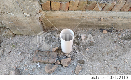 A white pvc pipe stands upright on a rough ground surface near a brick and concrete structure edge A white pvc pipe stands upright on a rough ground surface near a brick and concrete structure edge 132122110