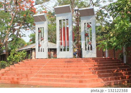A striking modern monument with three tall white pillars stands on a wide red brick staircase, surrounded by lush green trees 132122388
