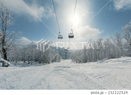 A ski lift moves upward through a winter landscape, surrounded by snow-covered trees and bright sunlight. 132122524