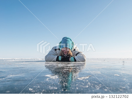 Young woman in winter attire enjoys lying on the clear, icy surface of Baikal Lake. 132122702