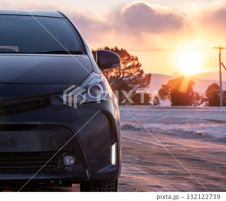 Black car parked on a snowy road during sunset near mountains reflecting golden light 132122739