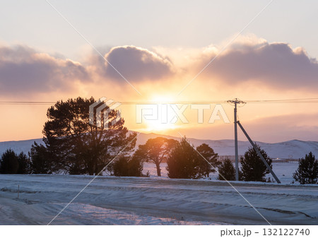Sunset over a frozen landscape with snow-covered ground and silhouettes of trees in the foreground Sunset over a frozen landscape with snow-covered ground and silhouettes of trees in the foreground 132122740