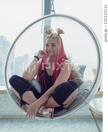Stylish woman with vibrant braids relaxes in a modern hanging chair against a city skyline backdrop Stylish woman with vibrant braids relaxes in a modern hanging chair against a city skyline backdrop 132123112