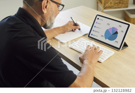 Senior man analyzing pie charts on tablet with keyboard at home office desk 132128982