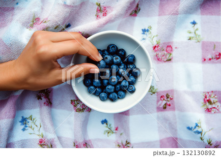 Hand picking fresh blueberries from white bowl on floral picnic blanket with soft pastel colors Hand picking fresh blueberries from white bowl on floral picnic blanket with soft pastel colors 132130892