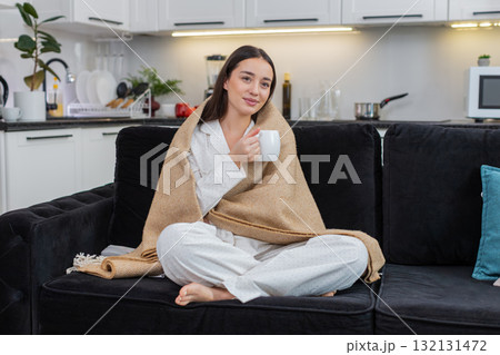 Young woman holds cup of tea wrapped in blanket relax calm cozy rest soft smile quiet evening hush 132131472