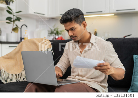 Indian man updates bill numbers in laptop spreadsheet checks docs and confirms monthly budget total Indian man updates bill numbers in laptop spreadsheet checks docs and confirms monthly budget total 132131501