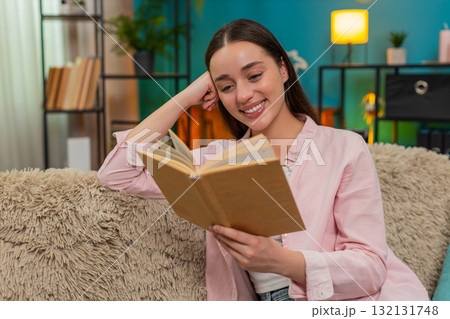 Young woman reading book on sofa turning pages slowly reflecting deeply on story absorbing meaning 132131748