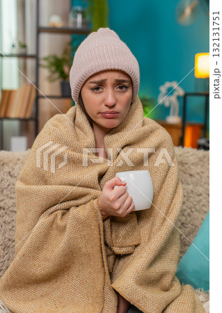 Young woman under blanket on sofa wearing wool hat drinking hot tea fighting bad cold with patience Young woman under blanket on sofa wearing wool hat drinking hot tea fighting bad cold with patience 132131751