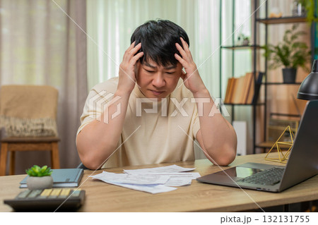Asian man holding head in stress, surrounded by unpaid bills on table, calculator and laptop nearby 132131755