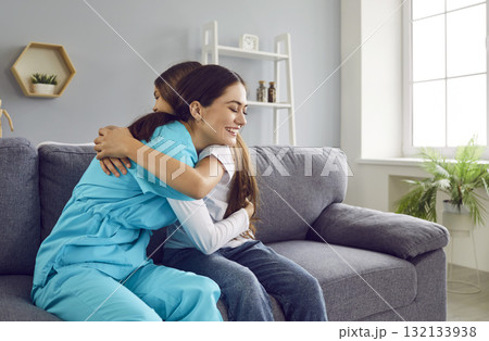 Portrait of a female friendly nurse with a child girl patient hugging sitting on sofa at home. 132133938