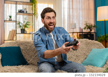 Young man intensely playing video game holding gamepad focused excited reacting home indoors sofa Young man intensely playing video game holding gamepad focused excited reacting home indoors sofa 132134029
