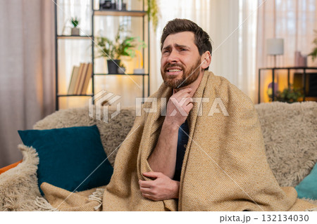 Young man sitting on the sofa at home, wrapped in a blanket, coughing, holding throat, feeling pain Young man sitting on the sofa at home, wrapped in a blanket, coughing, holding throat, feeling pain 132134030