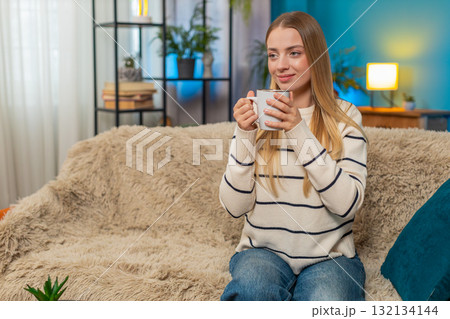 Caucasian woman enjoying hot tea on sofa holding cup both hands relaxing indoors calm comfortable Caucasian woman enjoying hot tea on sofa holding cup both hands relaxing indoors calm comfortable 132134144