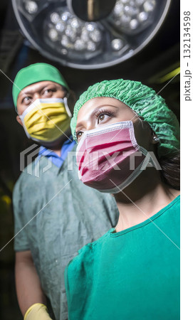Asian male surgeon and latina nurse preparing for complex operation, wearing protective masks, gloves and scrub attire. Bright operating room lights. 132134598