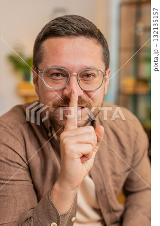Caucasian young businessman presses index finger to lips makes silence gesture at home office table 132135157