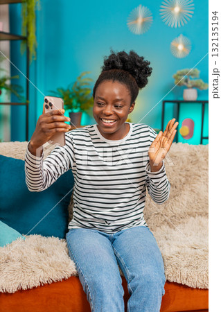 African American woman smiling waving smartphone hand gesture greeting happy indoors sofa African American woman smiling waving smartphone hand gesture greeting happy indoors sofa 132135194