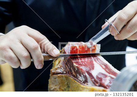 Professional chef slicing Iberico ham at gourmet kitchen in luxury restaurant. hands cutting cured meat delicately with knife and tongs, showing texture and craftsmanship in fine dining preparation. 132135201