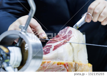 Professional chef slicing Iberico ham at gourmet kitchen in luxury restaurant. hands cutting cured meat delicately with knife and tongs, showing texture and craftsmanship in fine dining preparation. 132135203
