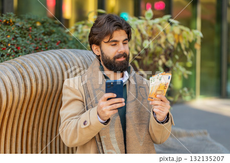 Happy rich Indian business man using smartphone and counting money euro on downtown city street 132135207