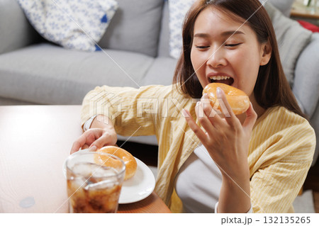 Happy young Asian woman enjoying a sweet glazed donut and iced cola at home, happy smiling in relaxation, showing simple joy and comfort food pleasure in a cozy lifestyle setting 132135265