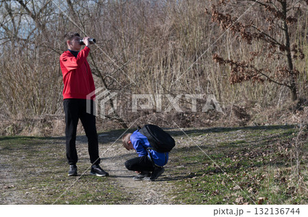 Two young people take a break during a hike, one drinks from a thermos, the other adjusts their backpack. Two young people take a break during a hike, one drinks from a thermos, the other adjusts their backpack. 132136744