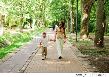 Front view of mother walking down the street with little son with sunglasses on sunny day in public green park. Front view of mother walking down the street with little son with sunglasses on sunny day in public green park. 132138645