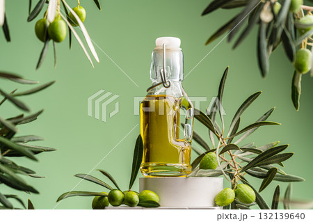 Glass bottle of olive oil with green olives and olive branches on a minimalist pedestal 132139640