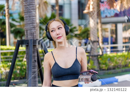 Exercise ready woman enjoys healthy sport workout, fit young person taking refreshing outdoor break from physical exercise, embracing wellness lifestyle with headphones, water and phone in urban park 132140122