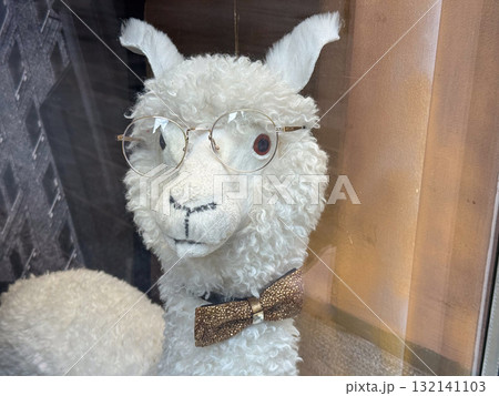 A white plush llama wearing glasses and a bow tie displayed in a shop window. Humor, personality, and charm reflected through playful modern decor. 132141103