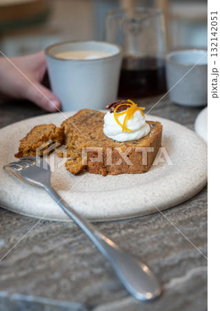 A slice of carrot cake with cream cheese frosting, pecan, and orange zest served with a cup of latte in a cozy cafe 132142051