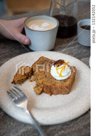 A slice of carrot cake with cream cheese frosting, pecan, and orange zest served with a cup of latte in a cozy cafe 132142052