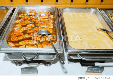 Buffet display featuring trays of sliced pork with sauce and cod fillet in a creamy sauce. A metal ladle rests in each tray, emphasizing a self-service setup 132142498