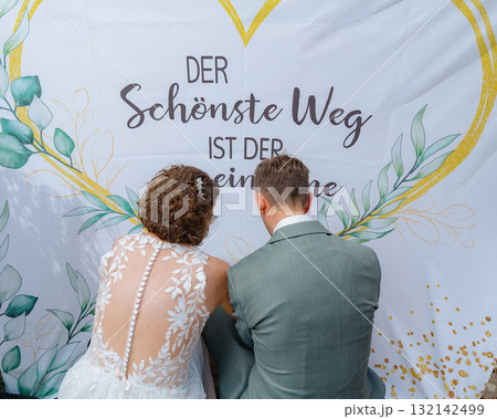 Bride and groom sit together signing a decorative backdrop on their wedding day. The text behind them suggests a romantic theme, enhancing the wedding ambiance and intimacy. Bride and groom sit together signing a decorative backdrop on their wedding day. The text behind them suggests a romantic theme, enhancing the wedding ambiance and intimacy. 132142499