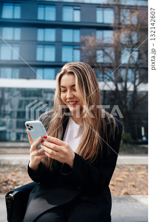 Young Woman Sitting Outdoors in Modern Urban Setting, Using Smartphone While Smiling During Sunny Day Young Woman Sitting Outdoors in Modern Urban Setting, Using Smartphone While Smiling During Sunny Day 132142607