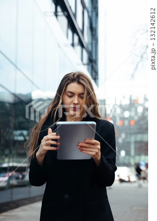 Young Woman Using a Tablet on a City Street While Dressed in a Formal Outfit During Daytime Near Modern Buildings and Glass Facades Young Woman Using a Tablet on a City Street While Dressed in a Formal Outfit During Daytime Near Modern Buildings and Glass Facades 132142612